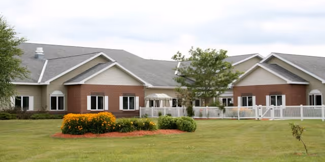 Exterior view of a single-story assisted living facility building with beige siding and red brick accents, white-framed windows, a white fence, and a landscaped lawn with bushes and flowers in the foreground under a cloudy sky.