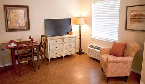A cozy living room area with a wooden floor, featuring a small dining table with two chairs, a flat-screen TV on a white dresser, a floor lamp, a window with blinds, and a patterned armchair with a red cushion.
