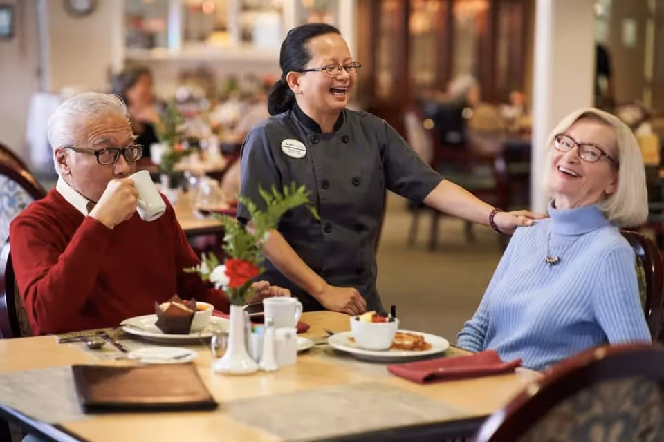 A smiling female staff member in a black uniform interacts warmly with two elderly residents seated at a dining table. The elderly man in a red sweater is drinking from a white cup, while the elderly woman in a light blue sweater is smiling. The table is set with plates of food, a flower vase, and dining utensils in a cozy dining room setting.