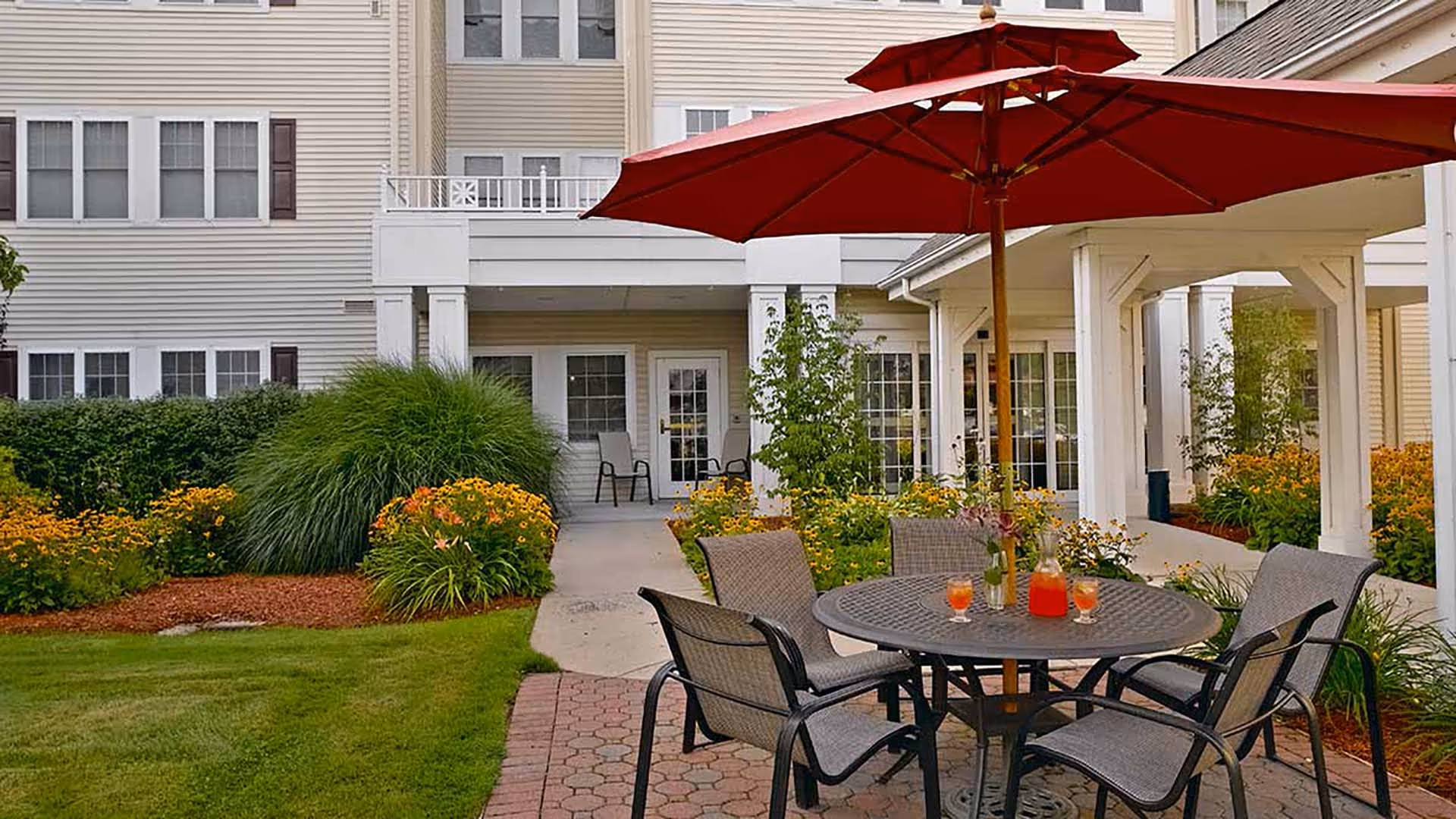 Outdoor patio area at a senior living facility with a round table, four chairs, and a large red umbrella. The table has a pitcher of red drink and three glasses. The patio is surrounded by green grass, flowering plants, and shrubs, with the building's beige exterior and windows in the background.