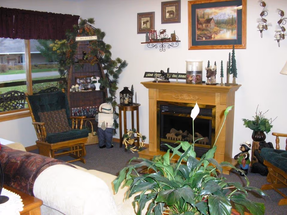 Cozy living room with a wooden rocking chair, a beige couch, a wooden fireplace mantel decorated with small figurines and a candle, a large green leafy plant in the foreground, a corner shelf with decorative items, and a window with a view of the outside.