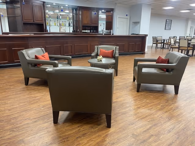 Four leather armchairs arranged around a small round table in a spacious room with a wooden bar and dining tables in the background.