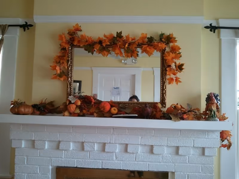 A white brick fireplace mantel decorated with autumn-themed garlands, pumpkins, and leaves. Above the mantel is a large ornate mirror framed with orange and red fall leaves. The wall behind is painted light yellow, and parts of white door frames and curtains are visible on either side.