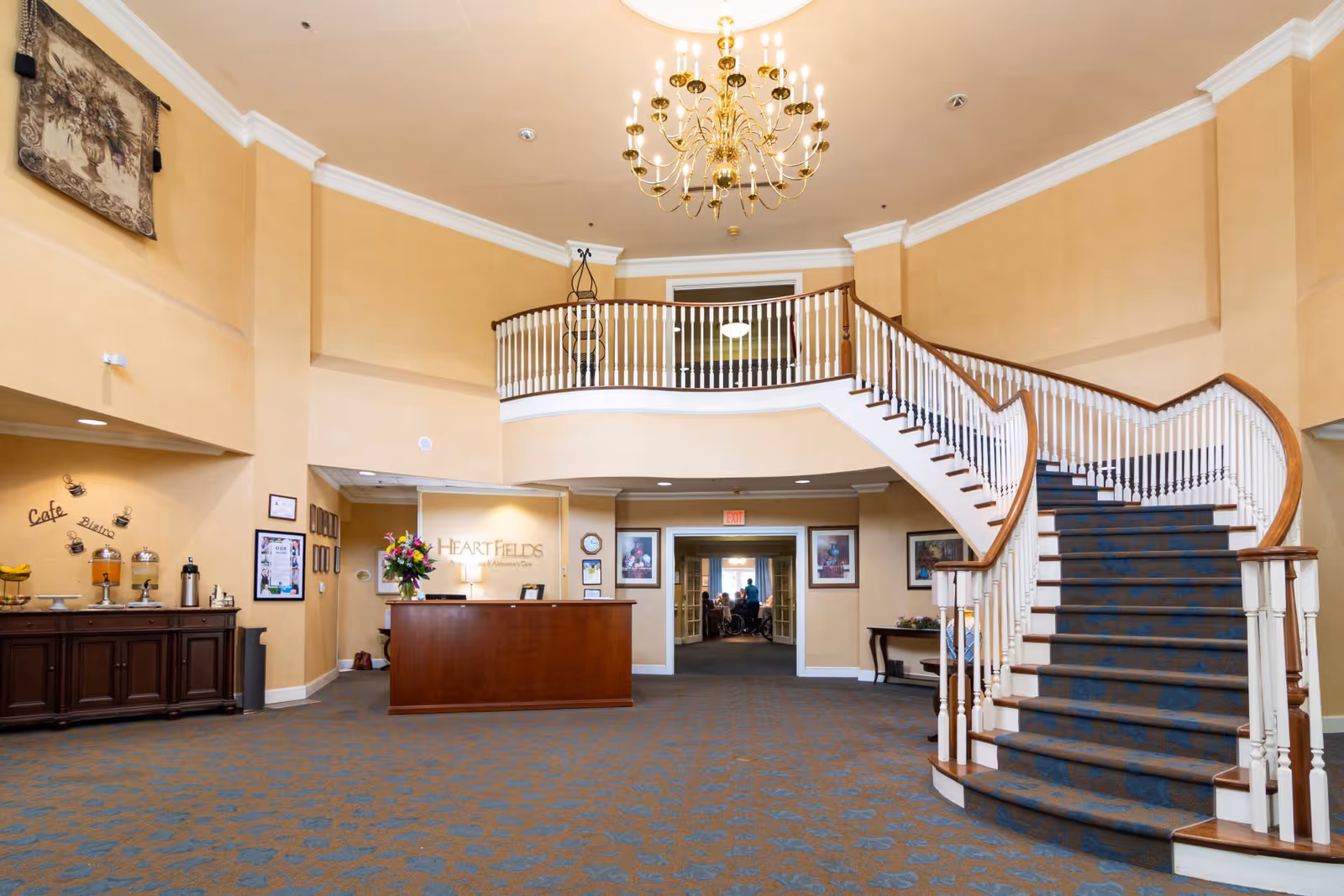 Spacious lobby area of HeartFields at Cary featuring a large curved staircase with a blue carpet runner, a chandelier hanging from the ceiling, a wooden reception desk with a flower arrangement, and a beverage station labeled 'Cafe Bistro' on the left side.