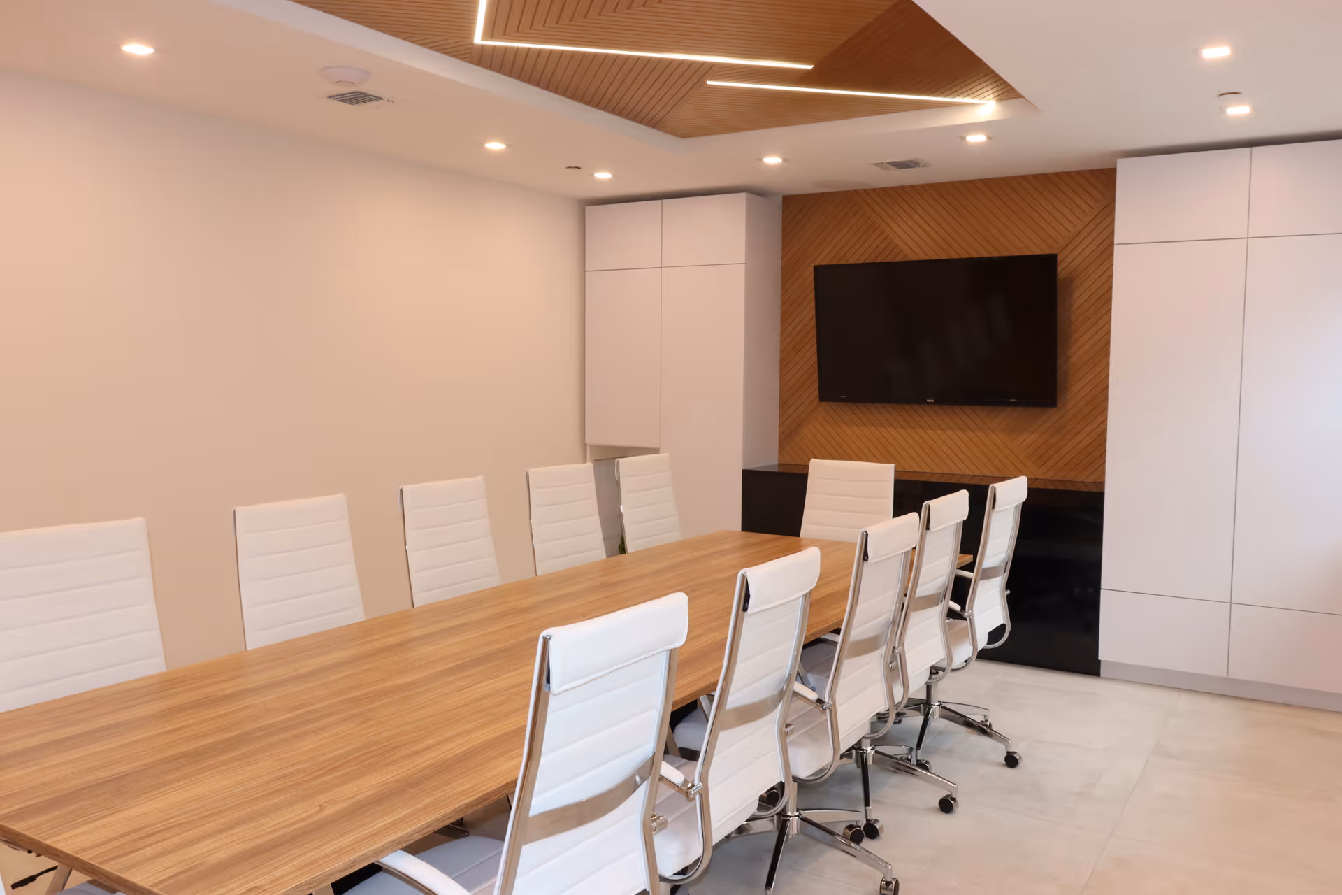 A modern conference room with a long wooden table surrounded by white office chairs on wheels. The room features a wooden accent wall with a mounted flat-screen TV, white cabinetry, and recessed ceiling lights.
