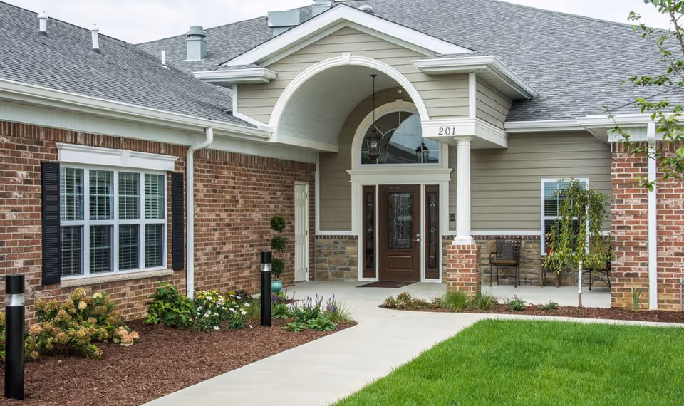 Front entrance of a residential building with a covered porch, brick and siding exterior, a brown door with glass panels, windows with white shutters, and landscaped garden beds along the walkway.