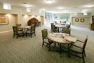 A spacious dining room with several round tables set with placemats, napkins, and utensils. The room has carpeted floors, large windows letting in natural light, and neutral-colored walls decorated with framed artwork and bulletin boards. Wooden chairs with cushioned seats surround the tables.