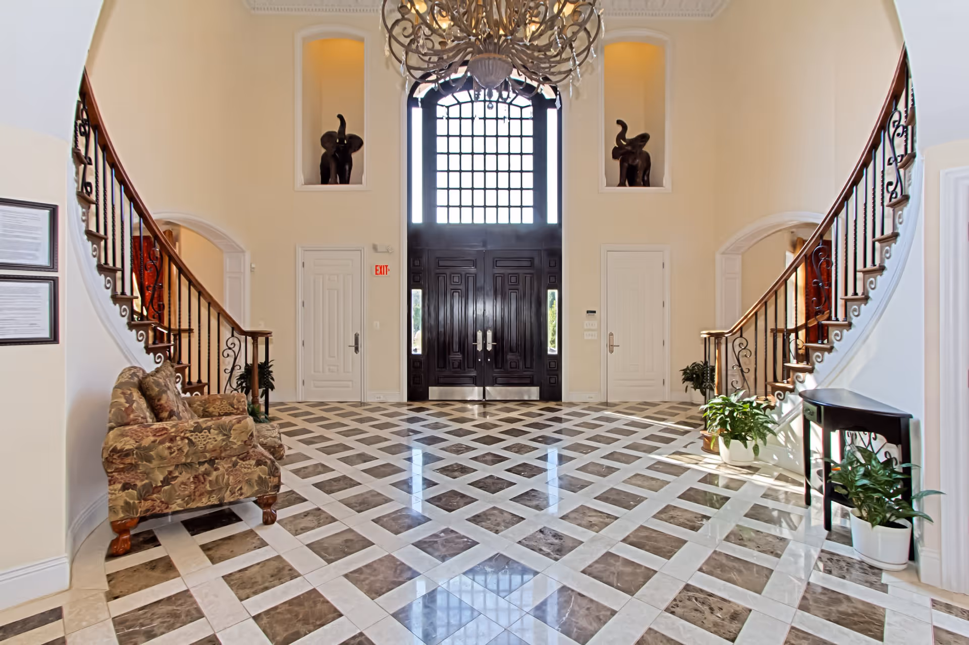 Spacious and elegant entryway with a large dark wooden double door featuring a grid window above it. The floor has a polished checkered pattern with brown and white tiles. Two curved staircases with wooden handrails and black iron balusters flank the sides. There are two floral upholstered armchairs on the left side and several green potted plants on the right side near a small black table. Two decorative elephant statues are placed in wall niches above the doors on either side of the main entrance. The walls are painted cream, and a large chandelier hangs from the ceiling.