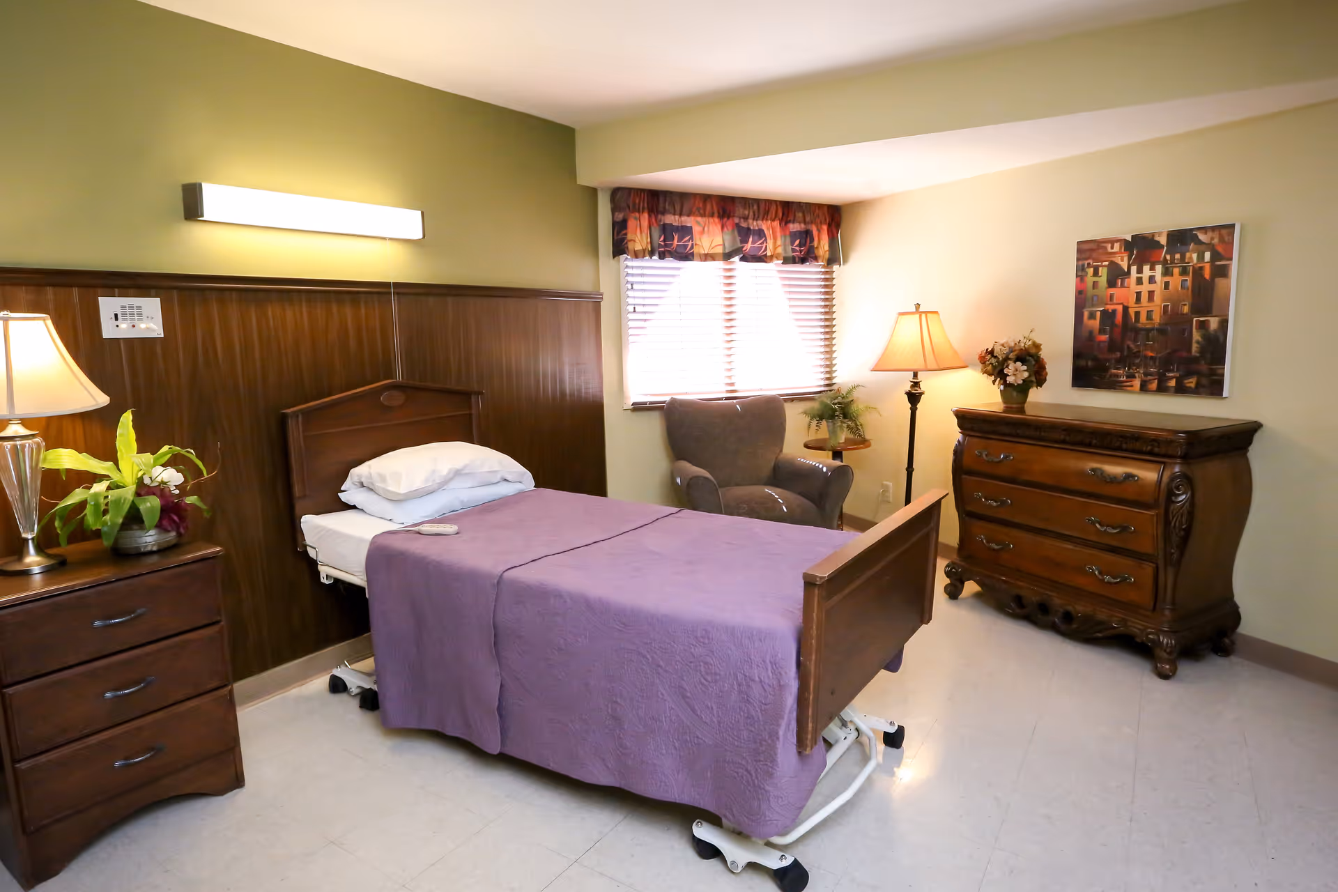 A neatly arranged bedroom in a senior living facility with a single bed covered in a purple blanket, a wooden headboard and footboard, a bedside table with a lamp and a plant, a comfortable armchair near a window with blinds and a valance, a floor lamp, a wooden dresser with a floral arrangement, and a framed painting on the wall.