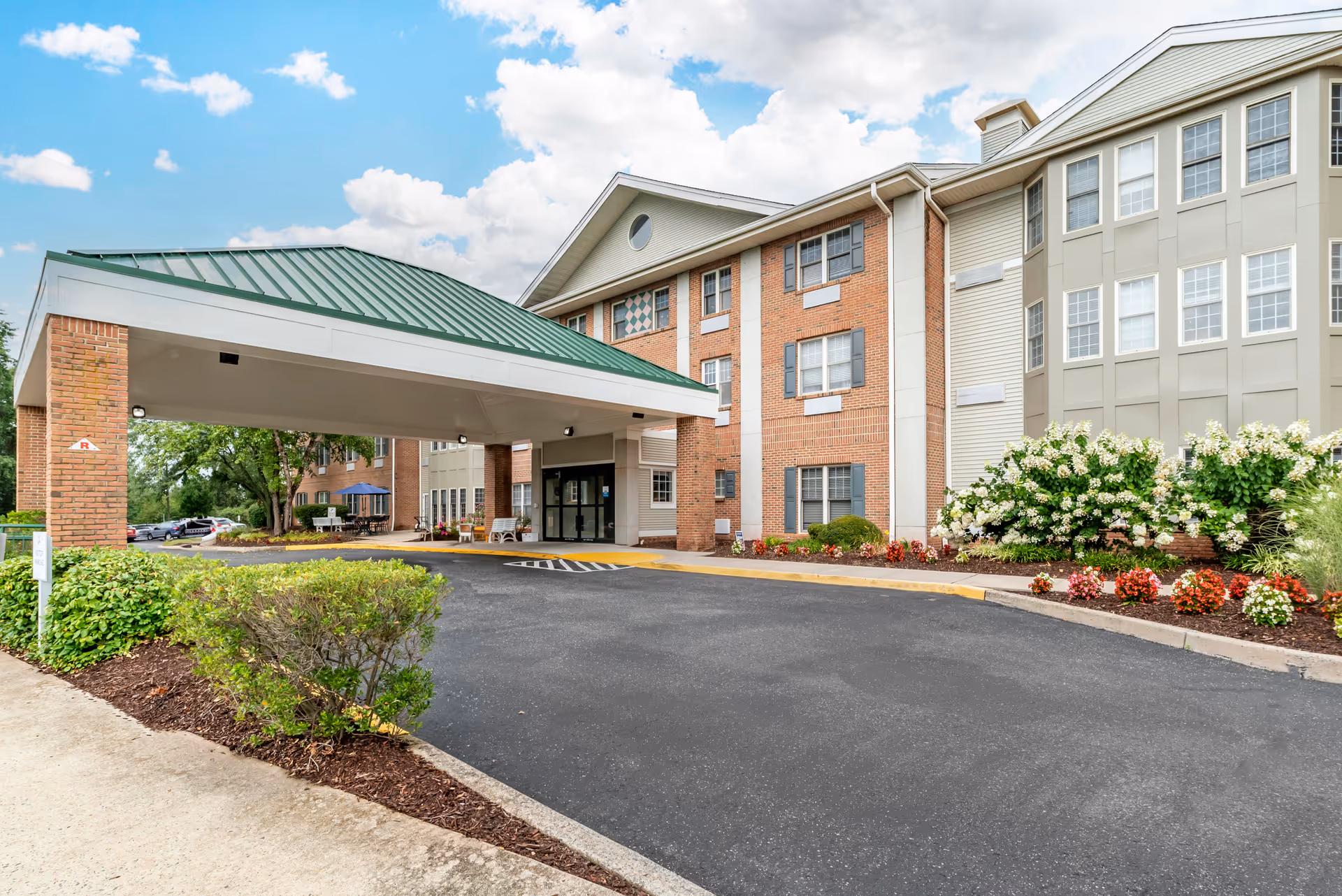 Exterior view of Brookdale Monroe senior living facility showing the main entrance with a covered drop-off area, brick and beige siding building, landscaped flower beds, and a clear blue sky with some clouds.