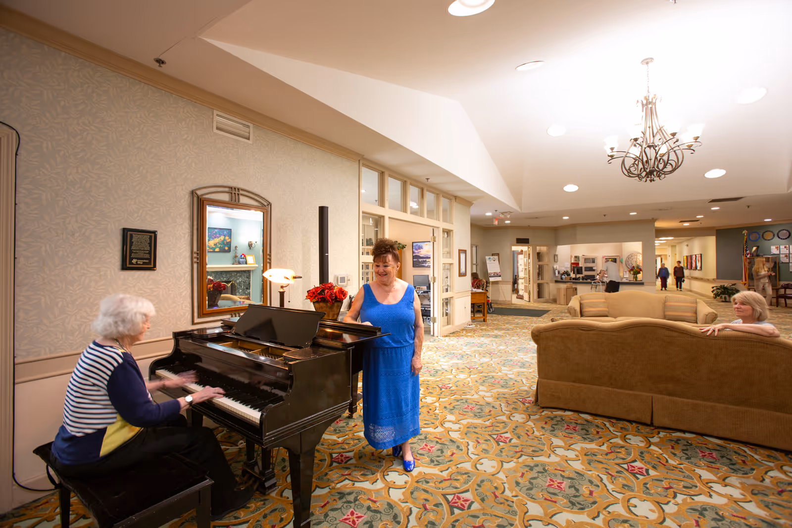 An elderly woman plays a black grand piano while another woman in a blue dress stands beside it smiling. Another woman is seated on a beige couch in a spacious, well-lit common area with patterned carpet, a chandelier, and several people walking in the background.
