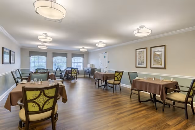 A dining room with several tables covered in brown tablecloths, each set with glasses and napkins. The room has wooden flooring, light-colored walls with a green lower half, and multiple windows letting in natural light. There are framed pictures on the walls and modern ceiling lights.