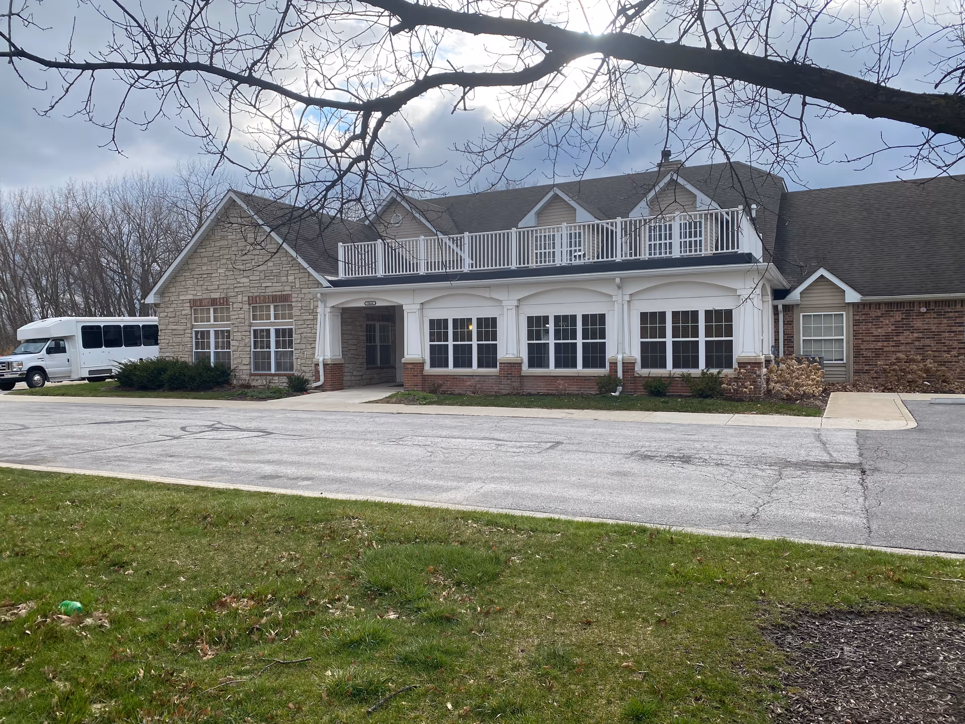 Front exterior of a brick-and-stone senior living building with large windows, white columns, a second-floor balcony, and a parked shuttle van.