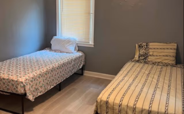 A small bedroom with two single beds placed against adjacent walls. One bed has a floral patterned bedspread and a white pillow, while the other bed has a striped bedspread with a matching pillow. There is a window with closed blinds on the wall between the beds. The walls are painted gray and the floor is light-colored wood.