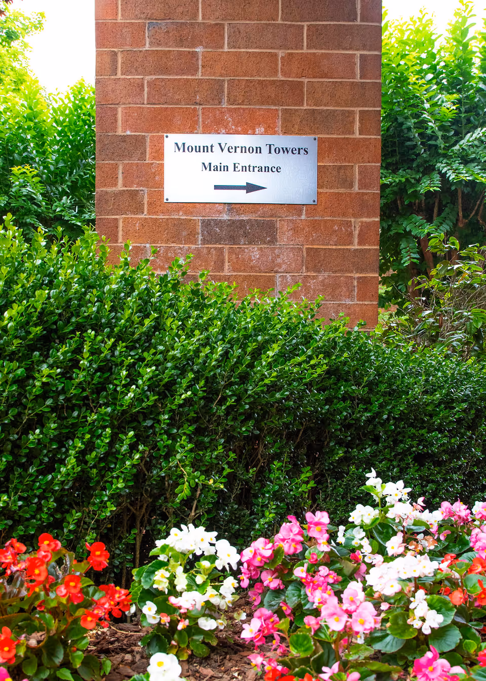 A brick wall with a metal sign that reads 'Mount Vernon Towers Main Entrance' with an arrow pointing to the right. In front of the wall, there are green bushes and colorful flowers including red, white, and pink blooms.