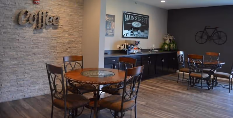 A cozy coffee area in an assisted living facility featuring round wooden tables with metal chairs, a countertop with coffee supplies, and wall decorations including a metal 'Coffee' sign and a framed 'Main Street Cafe' picture.