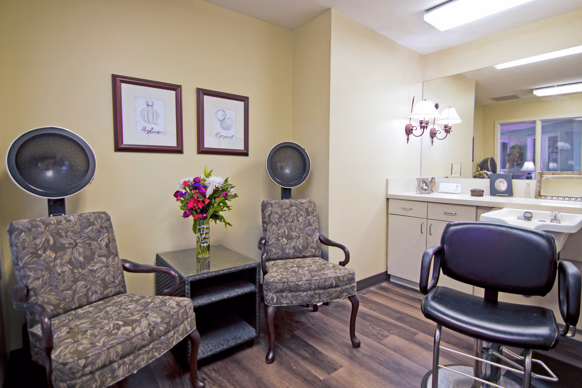 Interior view of a salon area with two vintage-style hair dryer chairs with floral upholstery, a small table with a vase of colorful flowers between them, a black salon chair in front of a sink and counter with cabinets, a large mirror, and wall-mounted light fixtures.