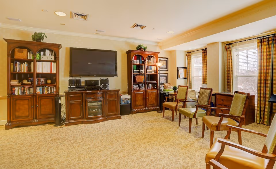 A cozy living room area with beige patterned carpet, two wooden bookshelves filled with books and decorative items, a flat-screen TV mounted on the wall above a wooden cabinet, and several wooden chairs with cushioned seats arranged around the room. The room has large windows with plaid curtains allowing natural light to enter.