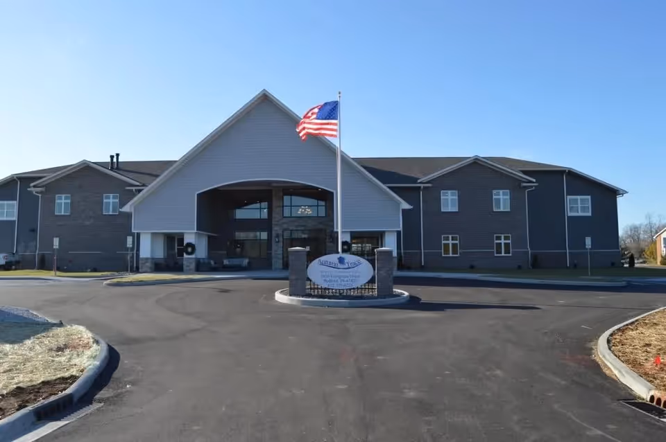 Front exterior view of Autumn Trace Bedford Assisted Living facility with a large entrance, an American flag on a flagpole, and a sign displaying the facility's name. The building is two stories with a combination of brick and siding under a clear blue sky.