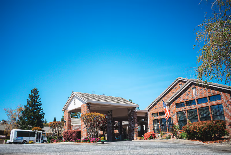 Front exterior of a brick assisted living facility with a covered entrance, American flag, landscaped shrubs and a shuttle van under a clear blue sky.