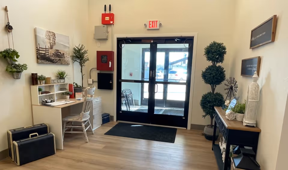 Interior view of an entrance area in The Kindred House facility featuring double glass doors with an exit sign above. To the left, there is a small desk with a chair, plants, and decorative items. On the right side, there is a console table with decorative plants and objects, and two framed quotes on the wall above it. The floor is light wood, and the walls are painted cream.