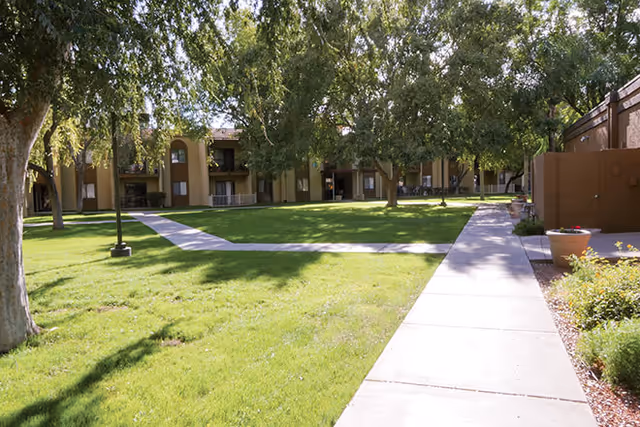 A grassy courtyard with trees, paved walkways, and a two-story residential building with balconies.