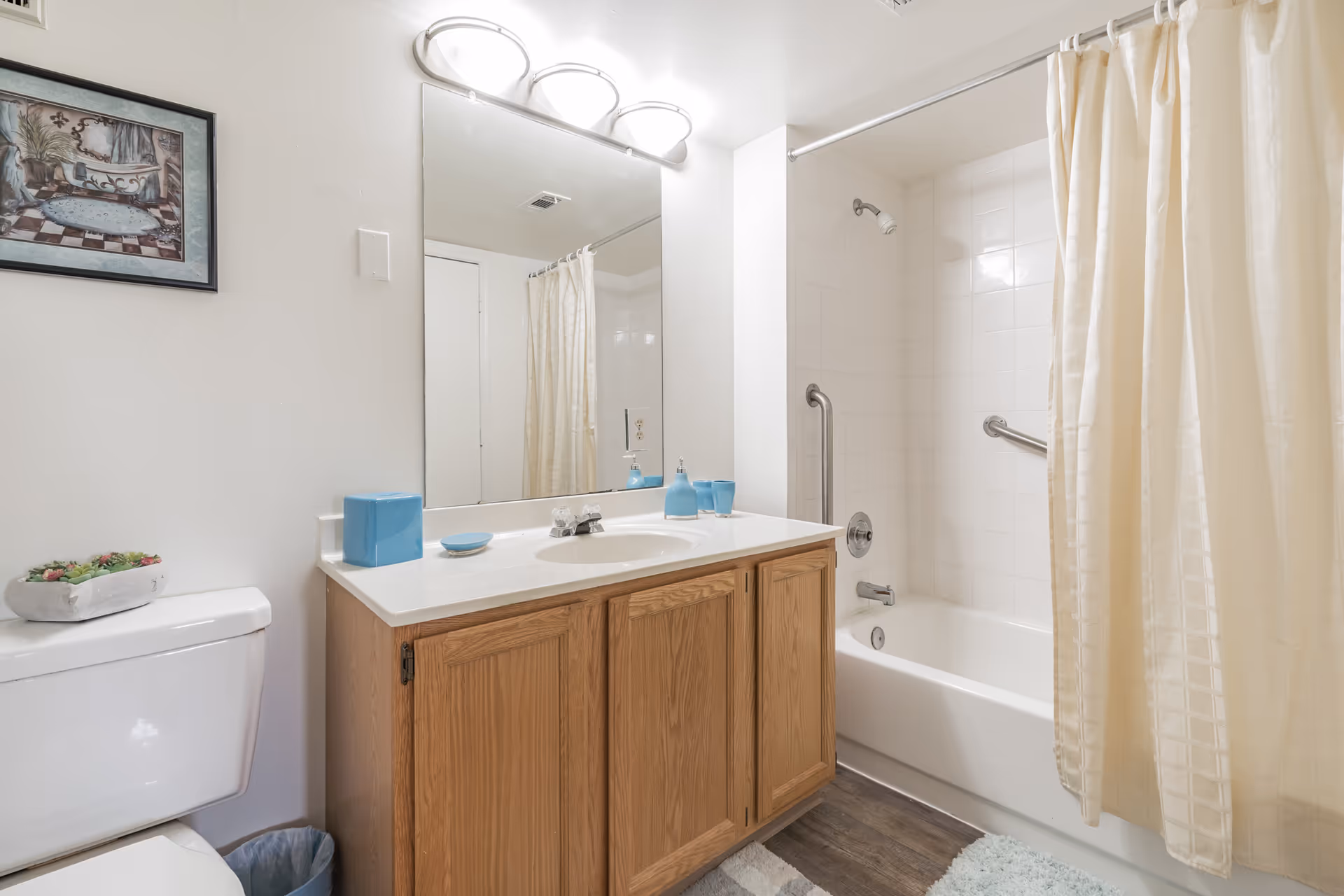 A clean bathroom featuring a white toilet with a small plant on top, a wooden vanity with a white countertop and sink, blue soap dispenser and cups, a large mirror with three overhead lights, and a bathtub with white tiled walls and a beige shower curtain.