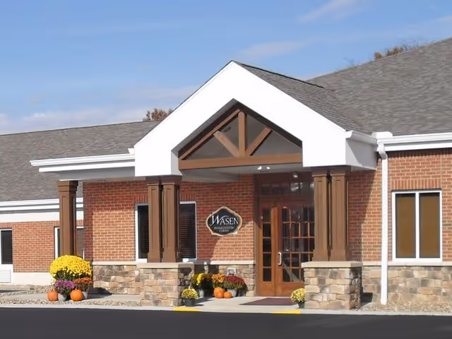 Entrance of a brick building with a peaked white roof supported by wooden columns and stone bases. There are flower pots with yellow and purple flowers and pumpkins near the entrance. A sign next to the door reads 'WASEN'.