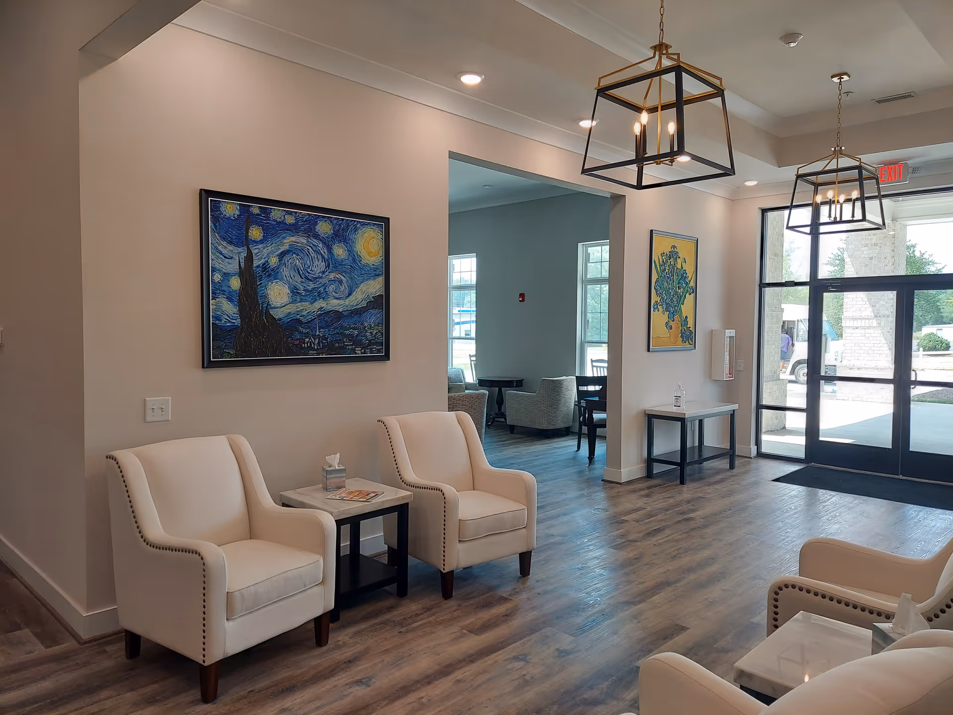 A bright and modern seating area in a senior living facility with cream-colored armchairs arranged around small tables. The room features wood flooring, large windows, and glass doors letting in natural light. Two framed paintings hang on the walls, including a reproduction of Van Gogh's Starry Night. Contemporary pendant lights hang from the ceiling.