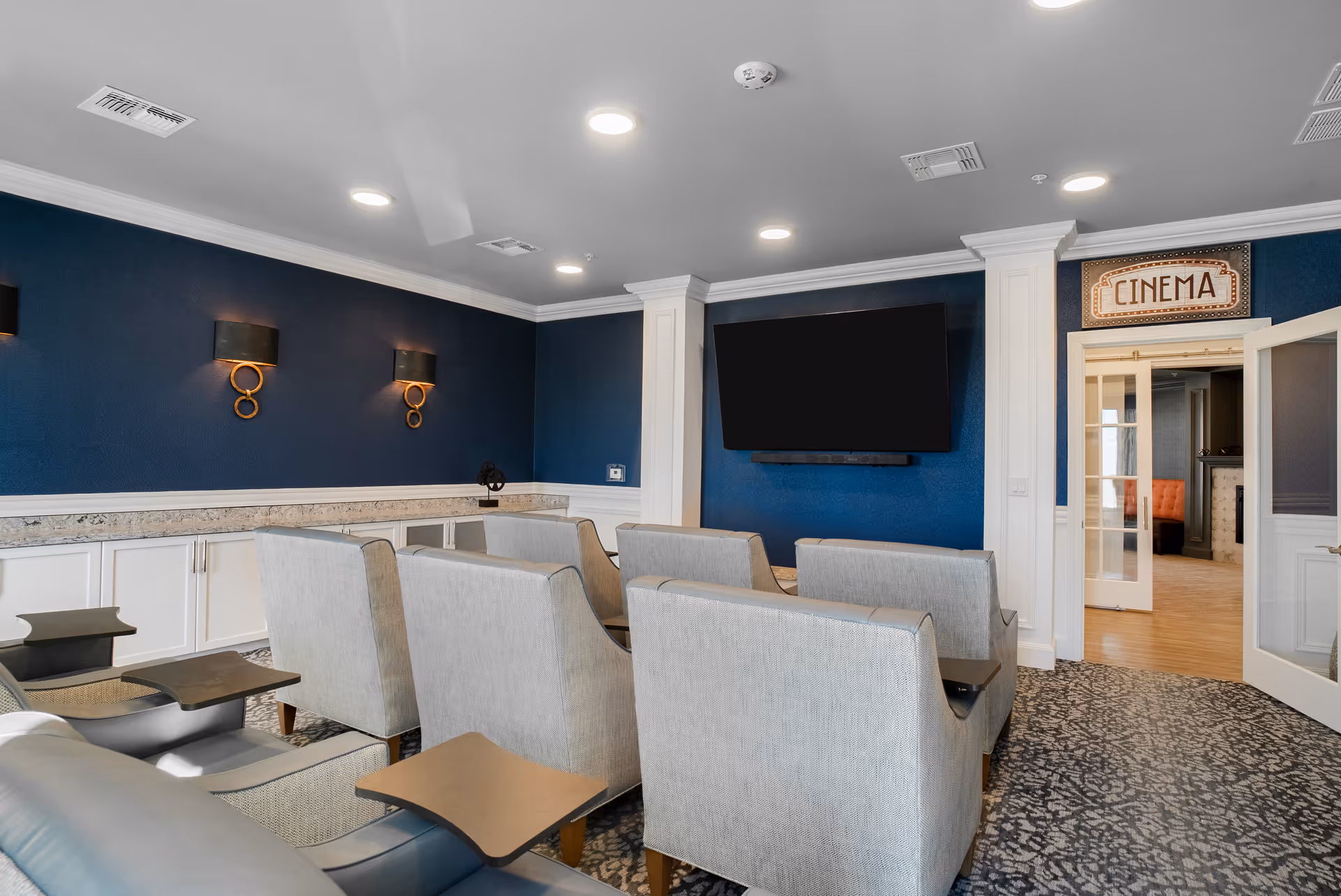 A small cinema room with several rows of comfortable gray chairs facing a large flat-screen TV mounted on a dark blue wall. The room has white trim and crown molding, recessed ceiling lights, and two decorative wall sconces. A sign above a glass door reads 'CINEMA,' and the door leads to another room with light wood flooring and an orange cushioned bench.