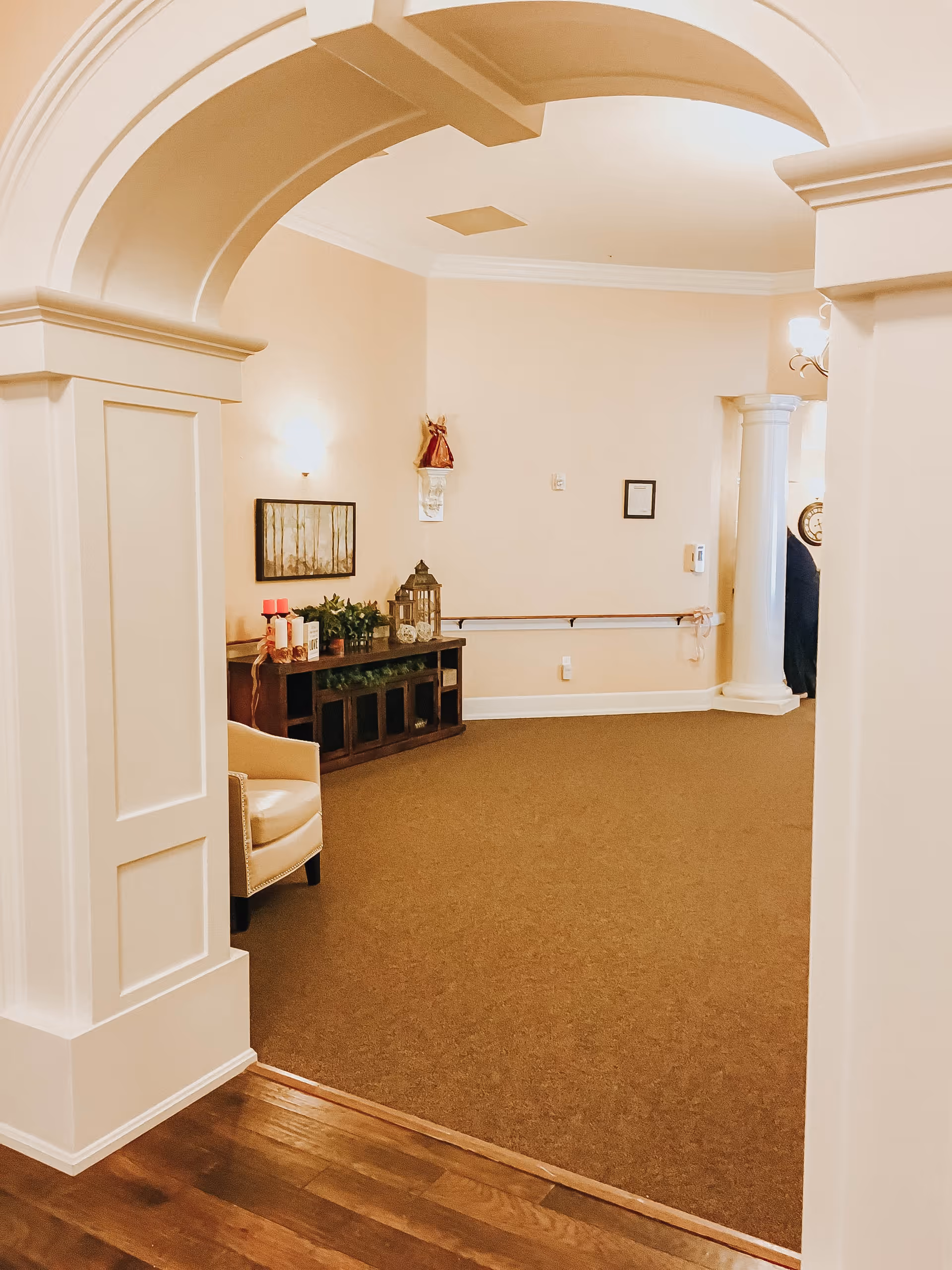 Interior view of a senior living facility hallway with beige walls, brown carpet, and wooden flooring. The space features an archway, a white armchair, a wooden console table decorated with plants, candles, and a lantern, and a white column in the background.