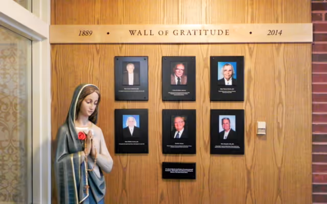A wooden wall display titled 'Wall of Gratitude 1889 - 2014' featuring six framed photographs of individuals with their names and titles beneath each photo. To the left of the display is a statue of a woman holding a rosary and a red rose, standing in a prayerful pose.