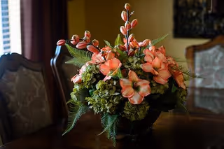 A floral arrangement with pink and green flowers and green leaves placed on a wooden table in a room with upholstered chairs and warm lighting.