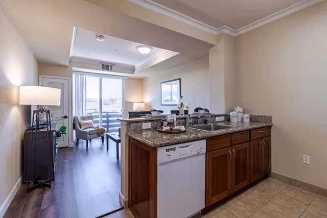 View of a modern kitchen area with granite countertops, wooden cabinets, and a dishwasher. The kitchen opens into a living room with a chair, coffee table, floor lamp, and sliding glass doors leading to a balcony with a scenic outdoor view.