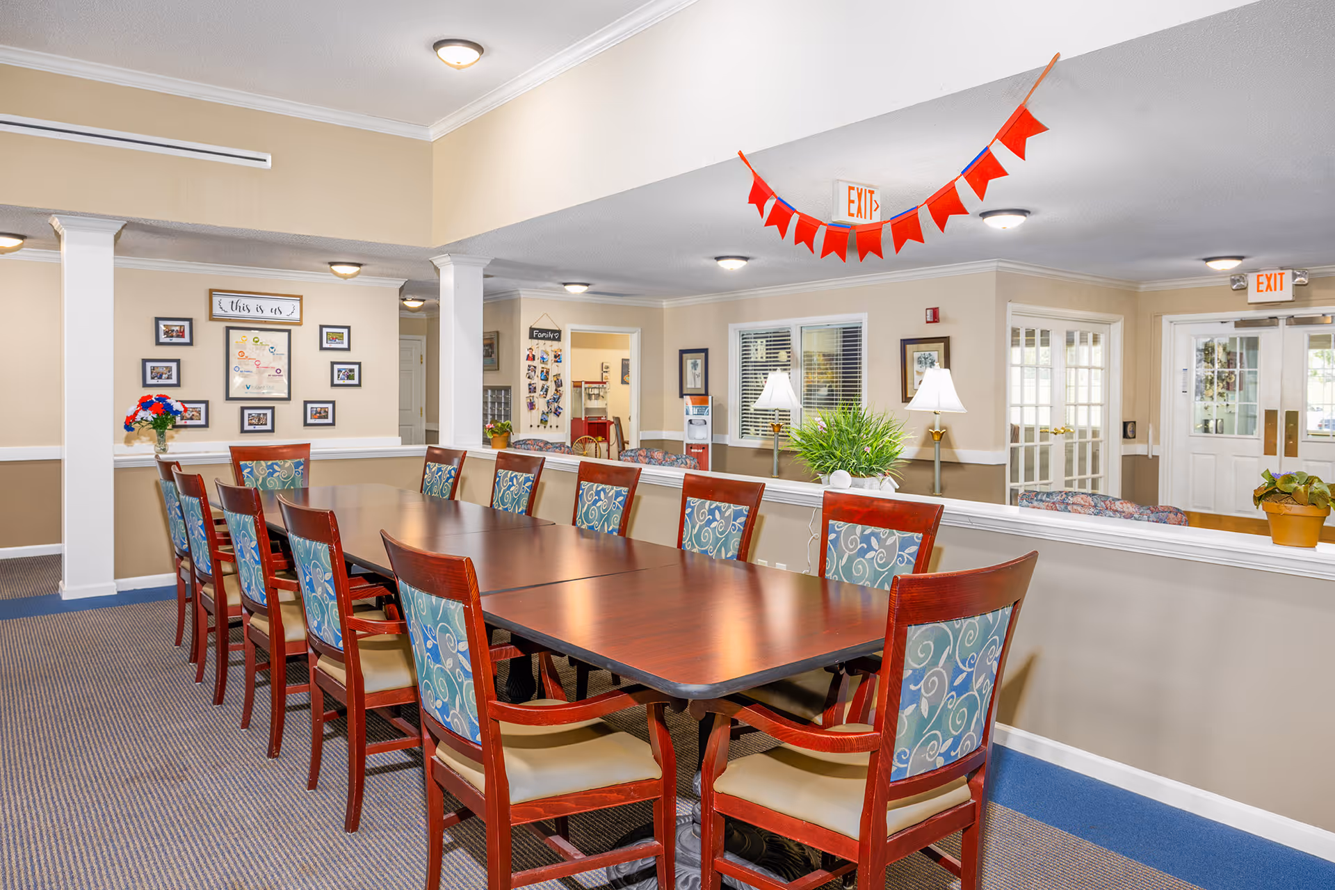 A senior living facility common area with a long wooden table surrounded by twelve chairs with patterned blue backs and beige seats. The room has beige walls, carpeted floors, and several ceiling lights. There are decorative plants on the half wall, framed pictures on the walls, and red triangular flags hanging from the ceiling near an exit sign. In the background, there is a water dispenser and a bulletin board with photos.