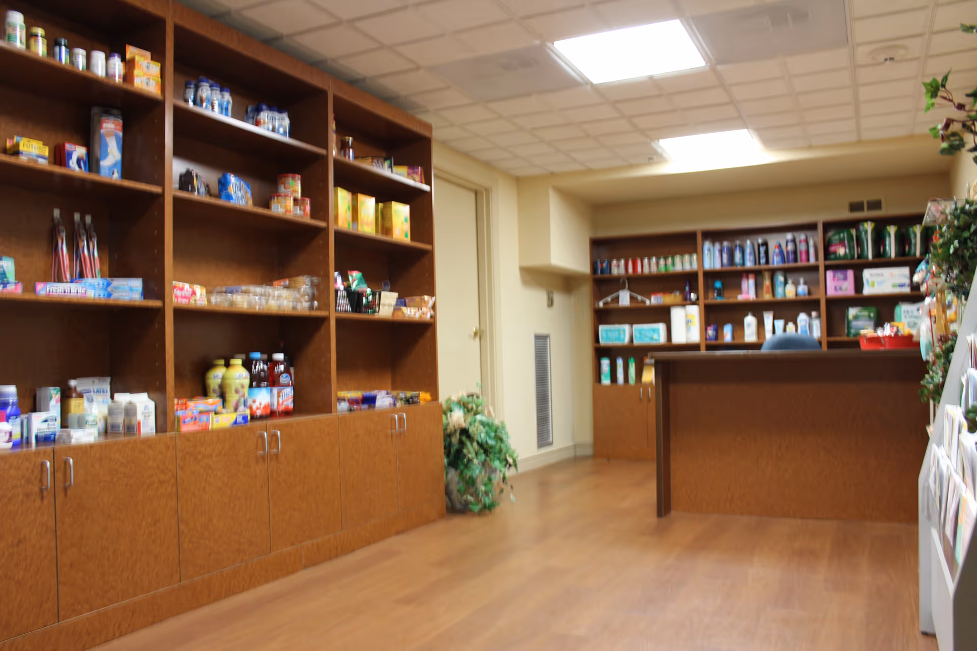 Interior view of a small convenience or supply store area within a senior living facility. Wooden shelves and cabinets line the walls, stocked with various products including snacks, beverages, and personal care items. There is a counter with additional shelves behind it, and a plant is placed near the corner. The room has a tiled ceiling with fluorescent lighting and a wood floor.