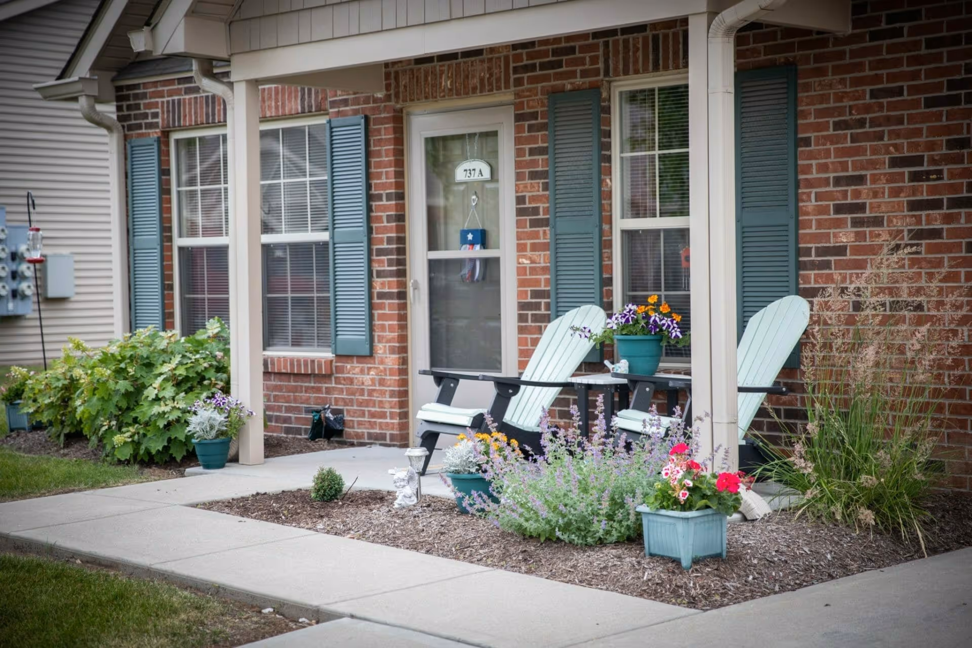 Front porch of a brick building with two light blue Adirondack chairs and a small table between them holding a flower pot. The porch is decorated with various potted plants and flowers, and there are blue shutters on the windows beside the door labeled 737 A.