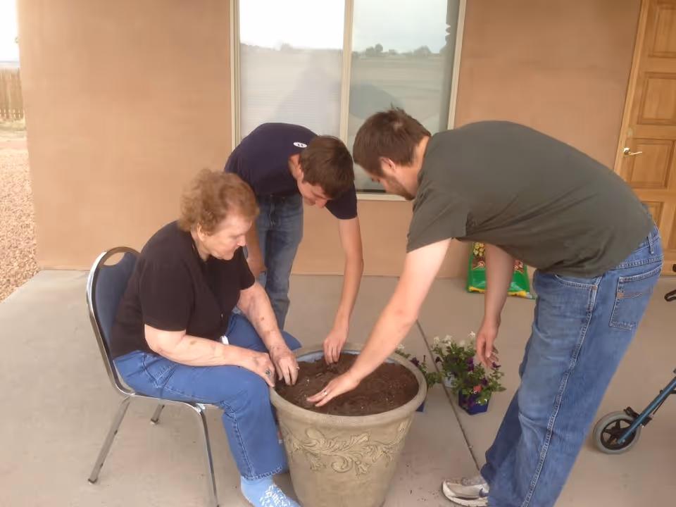 An elderly woman sitting on a chair and two younger men are planting flowers in a large decorative pot filled with soil on a concrete patio outside a building with a window and door in the background.