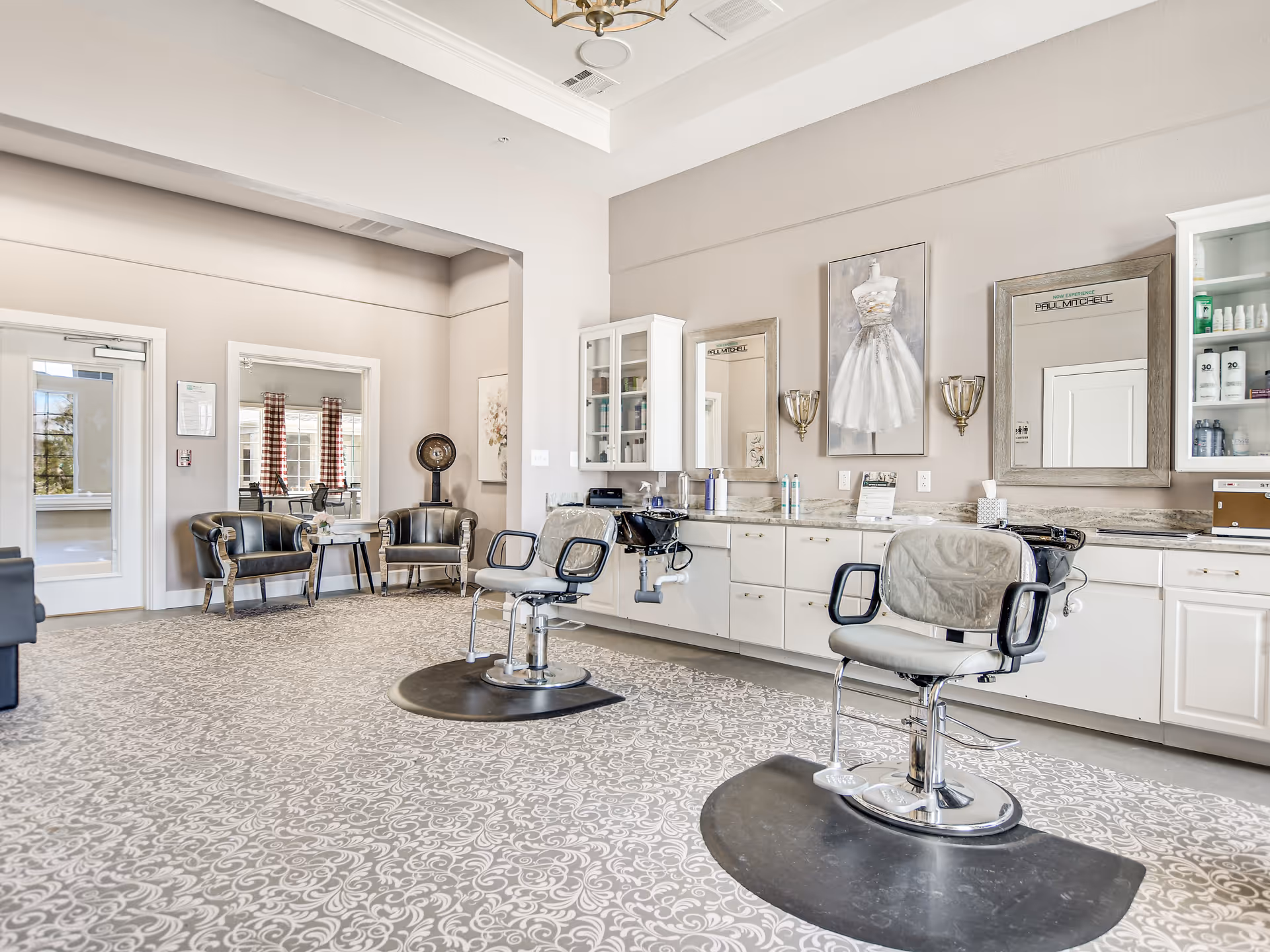 A bright and spacious salon area in a senior living facility with two salon chairs in front of large mirrors mounted on a wall with white cabinetry. The room features a patterned carpet, a painting of a white dress, and two black leather chairs with a small table near a door and window with red checkered curtains.