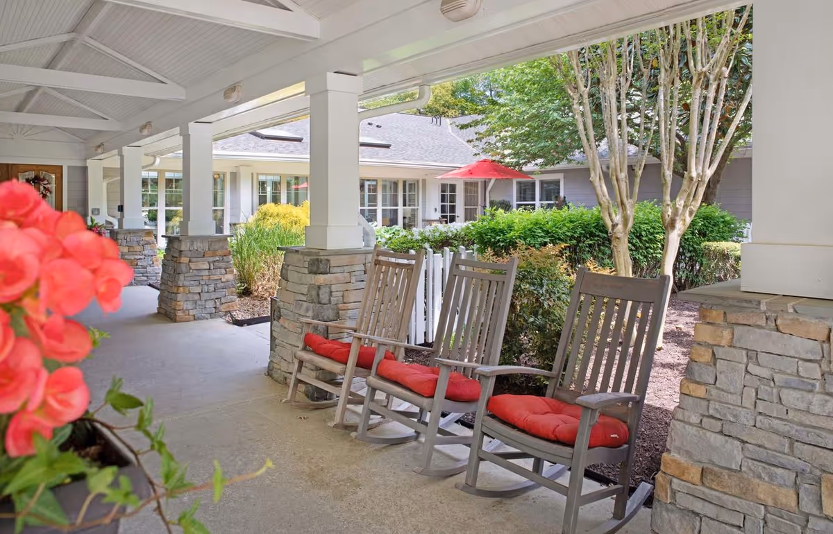 Covered front porch with three wooden rocking chairs with red cushions, stone columns, potted flowers, and a view of a courtyard and building with a red umbrella.