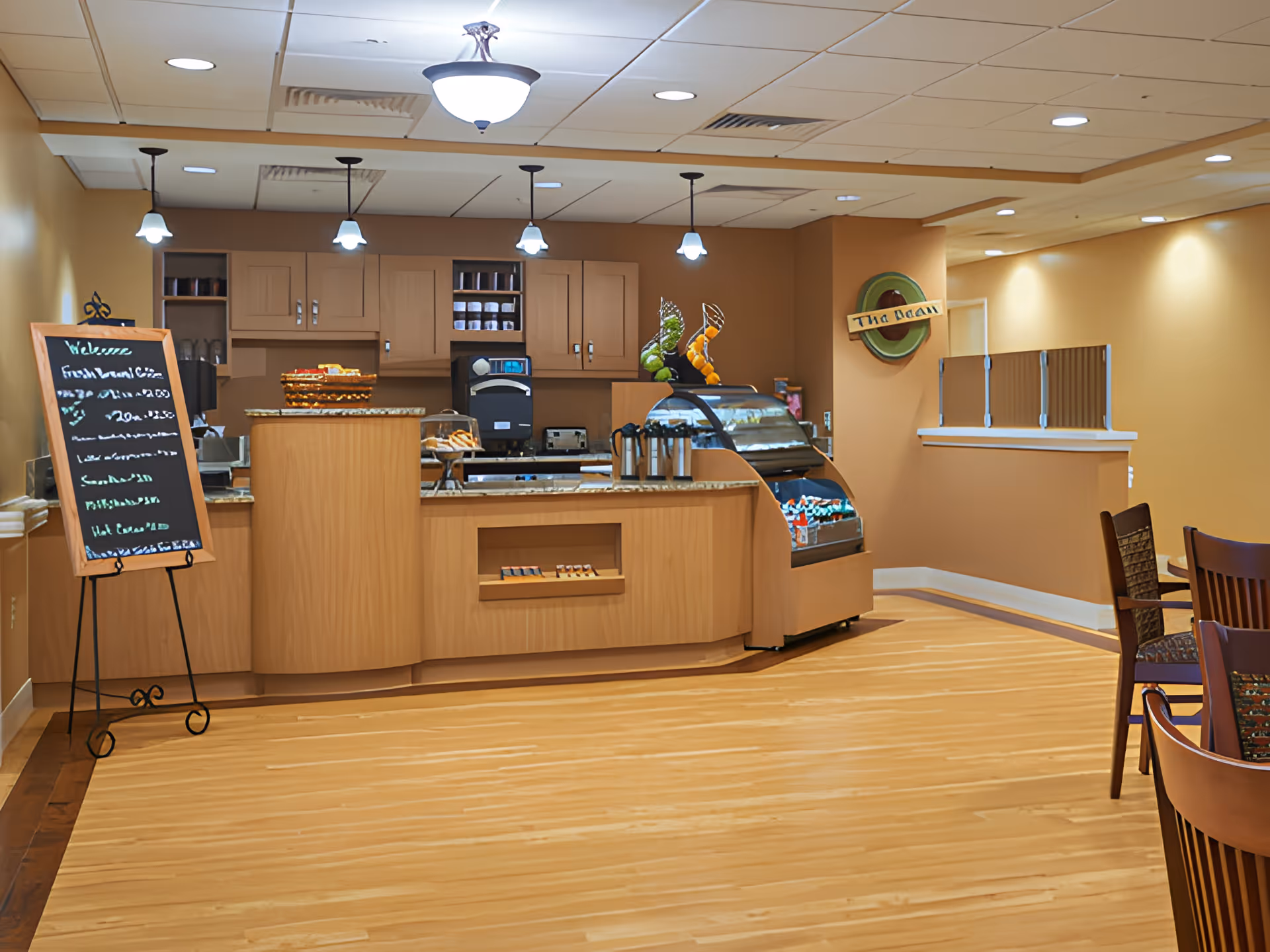Interior view of a small café or snack bar area with wooden cabinetry and counters, a display case with snacks, a chalkboard menu on a stand, pendant lights hanging from the ceiling, and a few wooden chairs and tables to the right. The walls are painted in warm beige tones.