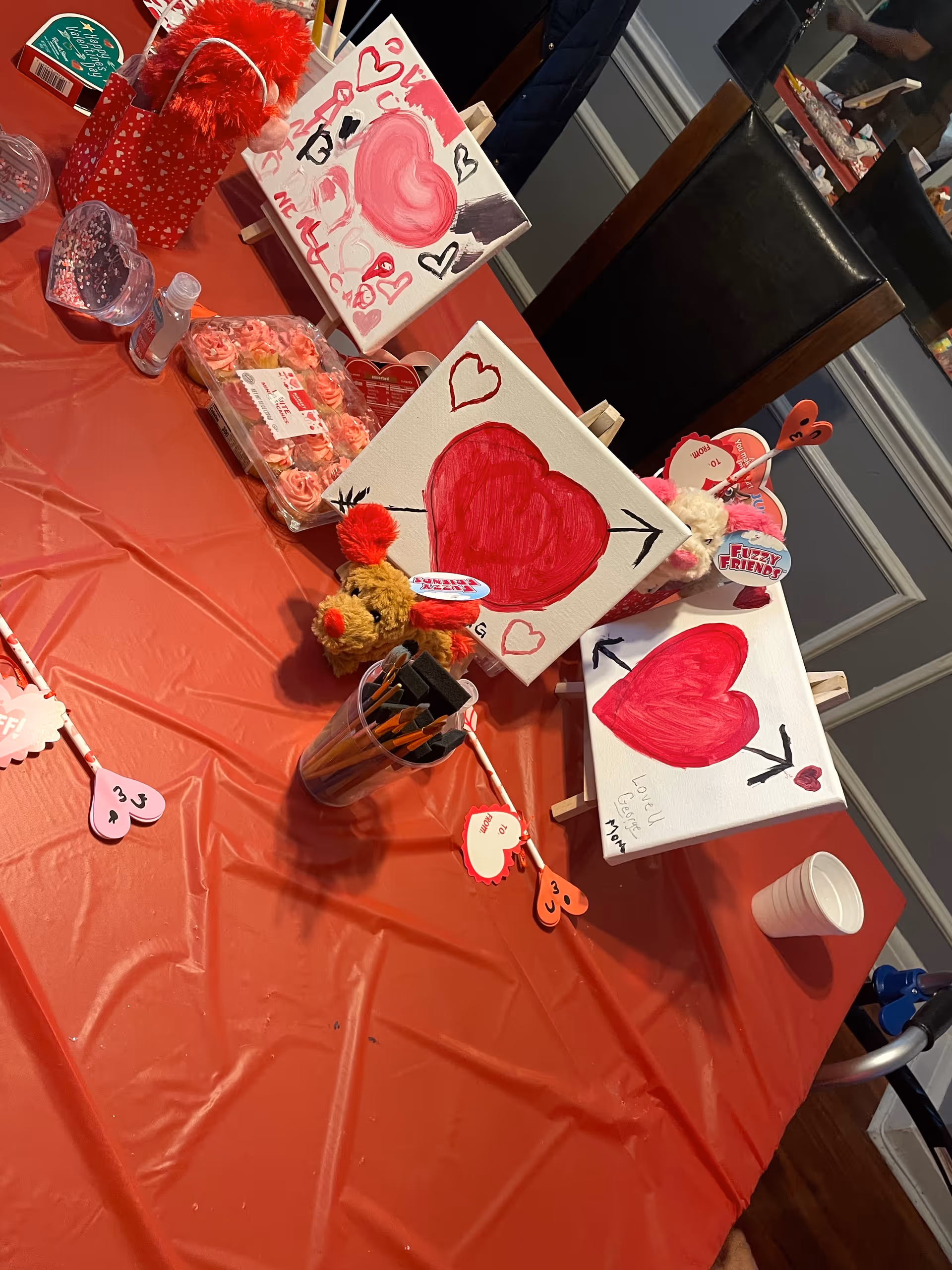 A table covered with a red plastic tablecloth displaying Valentine's Day themed crafts including paintings of red hearts with arrows, a small teddy bear with red pom-poms, heart-shaped decorations, a container with paintbrushes, and a tray of pink frosted cupcakes. The setting appears to be indoors with a mirror and chairs in the background.
