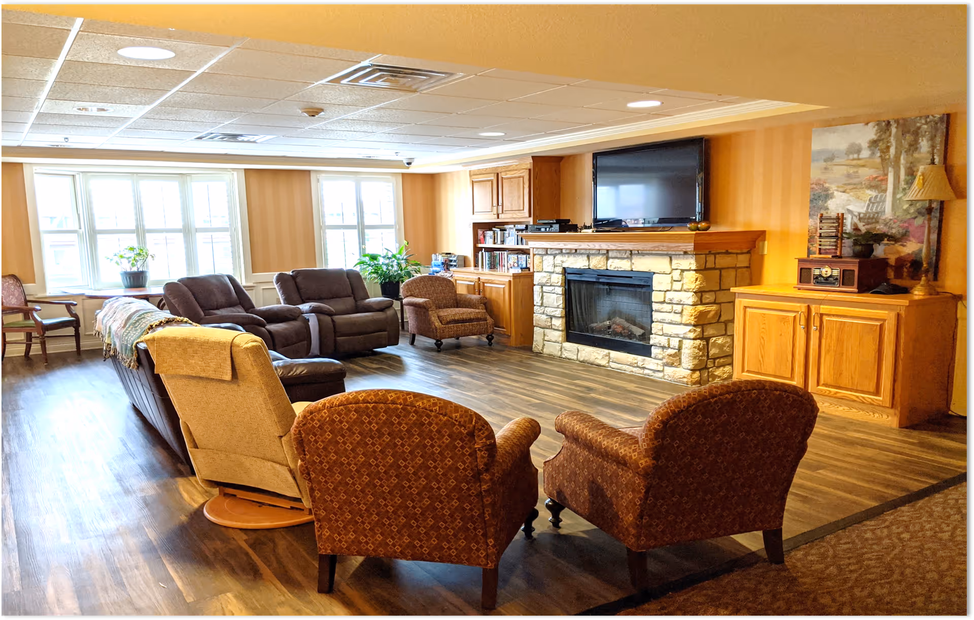 A cozy living room area with multiple upholstered chairs and recliners arranged around a stone fireplace with a mounted flat-screen TV above it. The room has wood flooring, built-in wooden cabinets, large windows letting in natural light, and decorative plants and artwork.