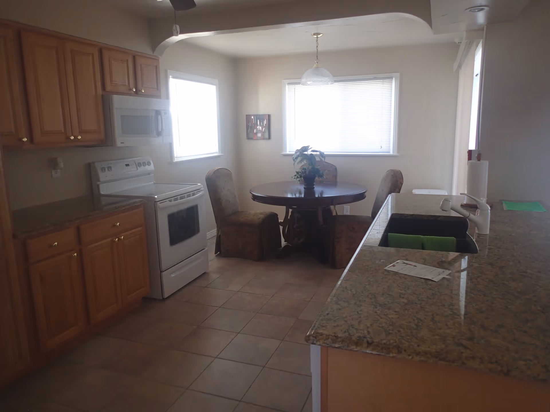 A kitchen and dining area with wooden cabinets, a white stove and microwave, a round dining table with four upholstered chairs, two windows with blinds, and a granite countertop with a sink and faucet.