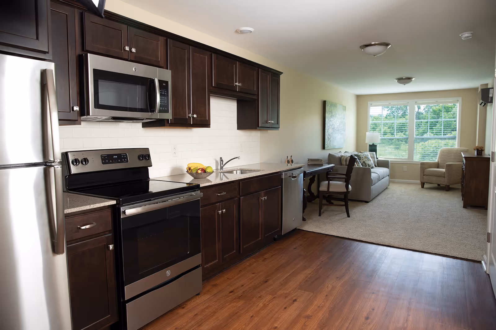 Interior view of a senior apartment featuring a kitchen with dark wood cabinets, stainless steel appliances including a refrigerator, microwave, stove, and dishwasher. The kitchen opens into a living area with a beige sofa, armchair, wooden side table, and large windows with white blinds letting in natural light.