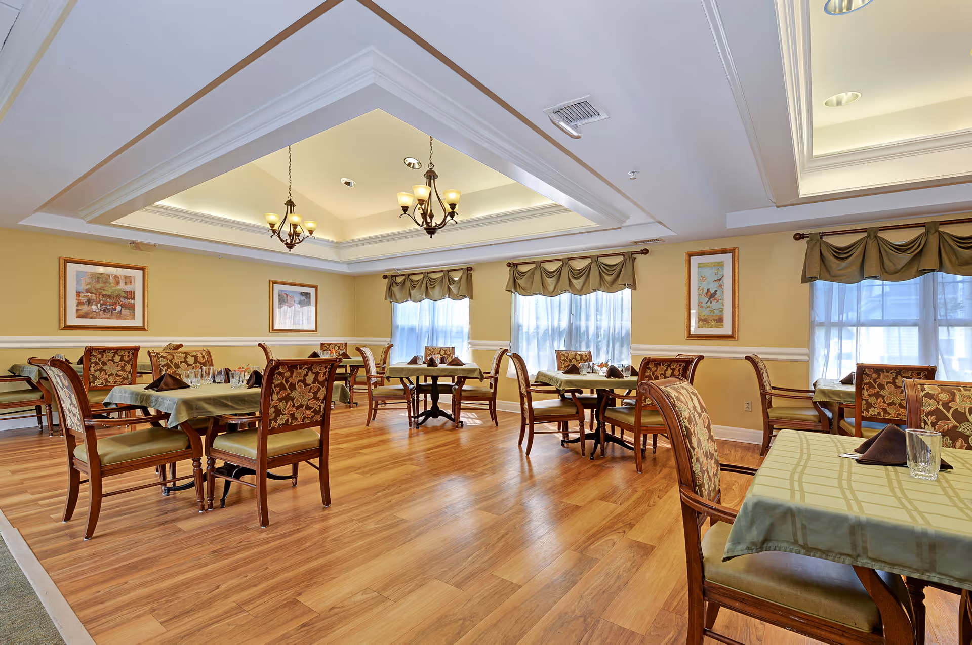 A bright dining room with wooden floors, several tables covered with green tablecloths, and chairs with floral upholstery. The room has large windows with green valances, framed artwork on the walls, and two chandeliers hanging from a recessed ceiling.