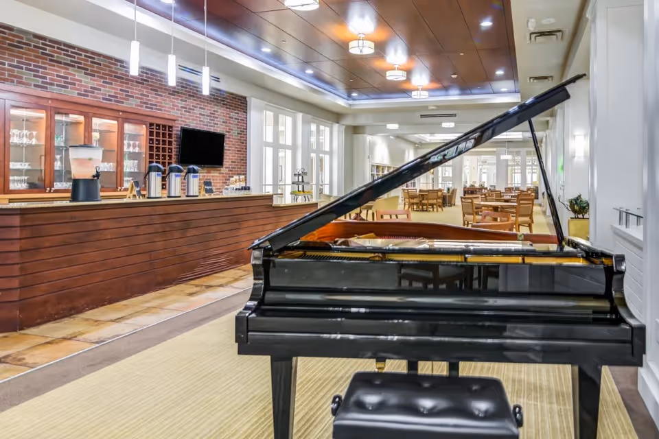 A grand piano in the foreground of a bright communal dining and lounge area with a wooden service counter and tables in the background.