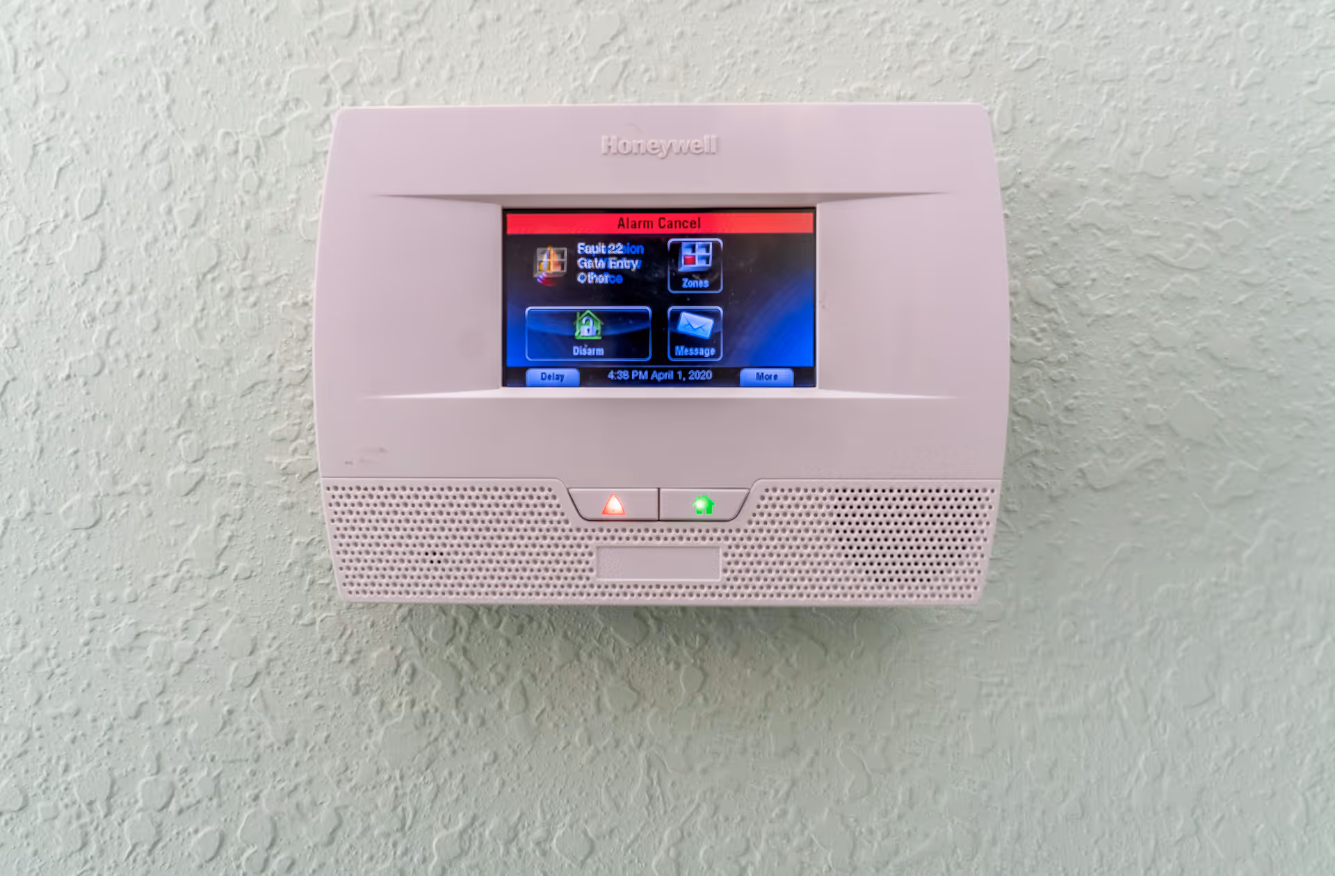 A Honeywell security system control panel mounted on a textured light green wall, displaying options such as Disarm, Zones, and Message on its screen.