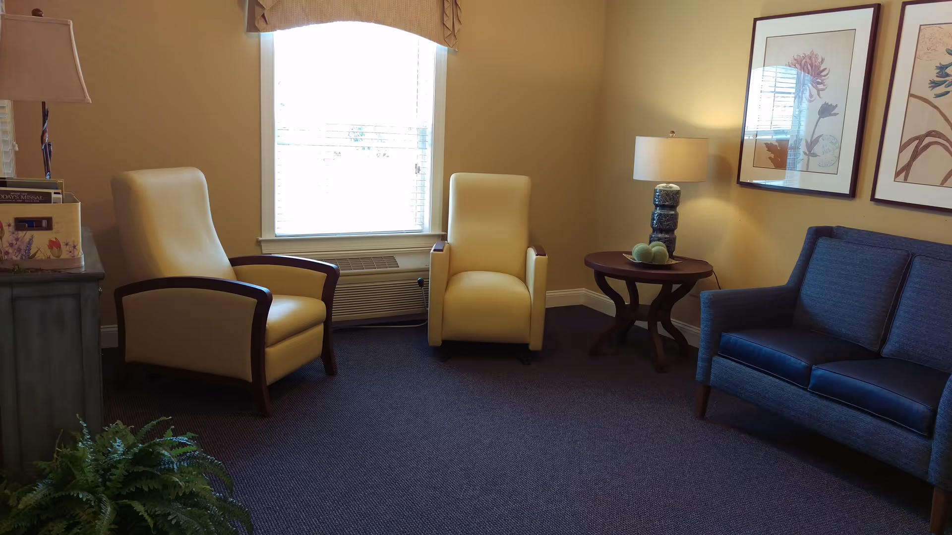 A cozy sitting area with two beige armchairs and a blue loveseat arranged around a small wooden side table with a lamp and decorative items. There is a window with blinds and a valance, framed botanical artwork on the walls, and a small green plant in the corner.