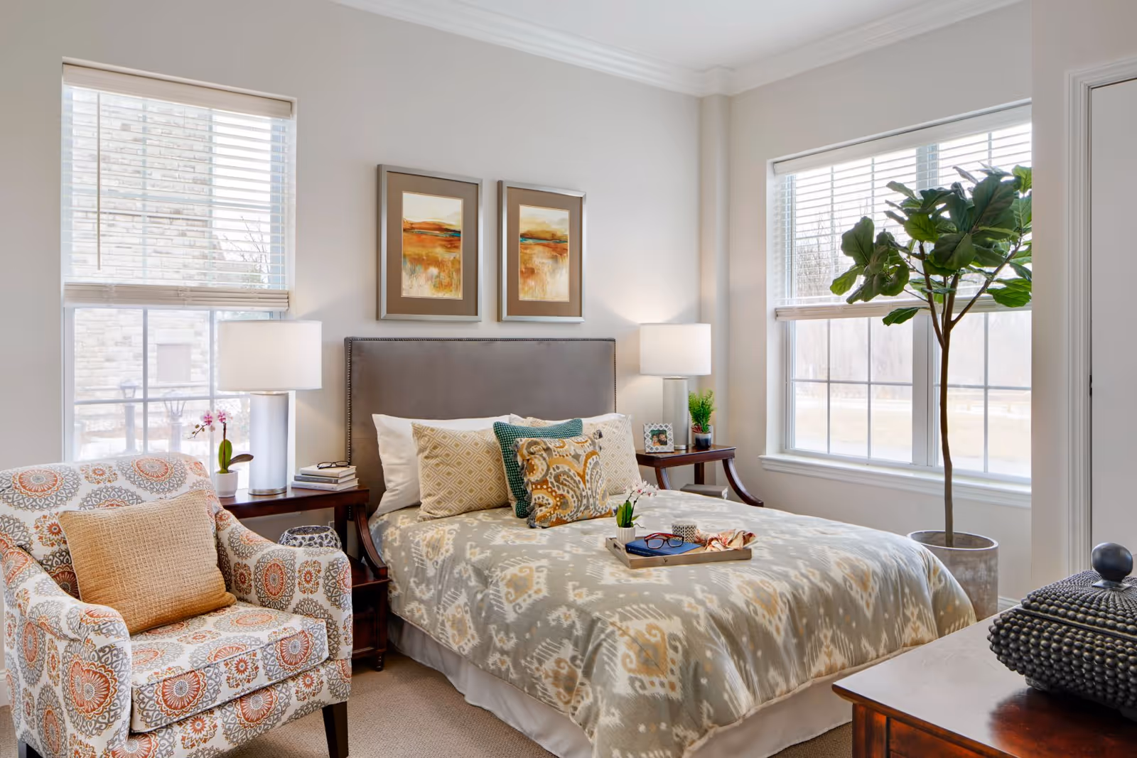 A cozy bedroom with a neatly made bed featuring patterned bedding and multiple decorative pillows. On either side of the bed are wooden nightstands with lamps, a small plant, and a framed photo. A patterned armchair with a beige cushion is positioned near a window with white blinds. Another large window with white blinds is next to a tall potted plant. Two framed landscape paintings hang above the bed.